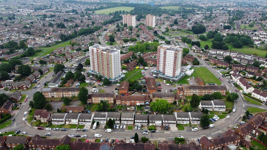 An aerial view of a residential area featuring a cluster of low-rise terraced houses arranged along curved streets, with front gardens and driveways. In the center, two multi-storey apartment buildings with light-colored façades and red accents are situated on a green patch, surrounded by parked cars and small trees. Behind the complex, the landscape extends into a suburban backdrop with additional housing and patches of greenery visible in the distance. The environment appears well-maintained, with neatly aligned vehicles and mature trees providing shade. The scene is evenly lit with natural daylight, highlighting the textures of the brickwork, roofing materials, and lush foliage. This image, associated with waste clearance services, illustrates a typical urban residential neighborhood potentially serviced by independent waste collection or rubbish removal companies such as Waste Clearance Harrow, especially relevant for managing waste from multi-unit and private properties in Harrow.