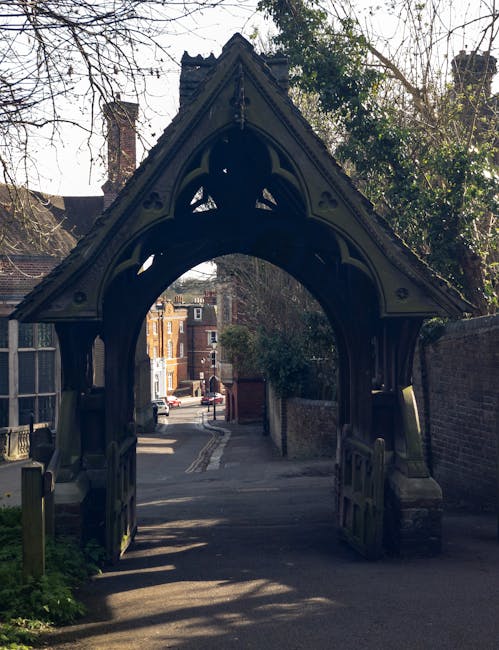 A historic, ornate black-painted metal archway with decorative Gothic Revival-style detailing, lining the entrance to a narrow alleyway or driveway. The archway features pointed arch shapes with intricate cut-out patterns and small floral motifs along the edges. In the background, there are residential buildings made of brick, with some windows and chimneys visible, along with trees casting shadows on the ground. The scene is captured during daylight with shadows indicating the sun's position, and the surrounding environment includes a brick wall on the right and greenery on the left. The alleyway behind the archway appears to lead to a residential street with parked cars and a few pedestrians, suggesting an urban setting where independent waste management or private rubbish removal might occur, supported by companies like Waste Clearance Harrow. The overall atmosphere is quiet and suburban, with the archway acting as a distinctive entrance to the private property or service area.