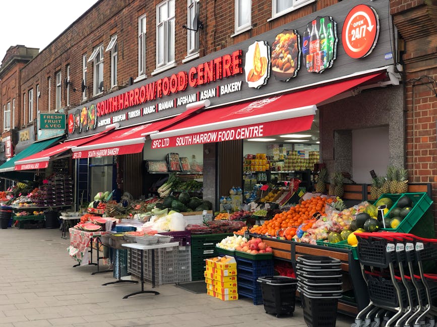 The image depicts the exterior of a South Harrow food store with a prominent red and white striped awning displaying the store's name and various types of food such as bakery items, Asian and Afghan products, and beverages. In front of the store, the pavement is lined with multiple open-fronted crates and containers filled with fresh fruits and vegetables including oranges, watermelons, apples, and other produce, some in plastic bags. There are additional display tables with packaged goods, while the storefront has signage indicating it operates 24/7. To the left, there are smaller green and black plastic crates stacked beside a small table with more produce, and to the far left, a green awning covers a stock of flowers and plants for sale. The multi-story brick residential building above features several white-framed windows, with some opened, under a cloudy sky. The environment suggests a busy street scene with active retail activity and potential waste from daily customer shopping, which Waste Clearance Harrow could assist in managing through private, on-site clearance of surplus items or rubbish related to grocery and retail waste handling.