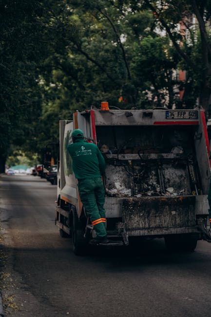 A waste collection worker dressed in a green uniform and safety helmet is standing on the pavement beside a small, commercial waste collection vehicle with a predominantly white body and red accents at the rear. The vehicle's rear hatch is open, revealing a blackened, soot-stained compartment used for loading rubbish. The worker appears to be inspecting or preparing the load for disposal, and the vehicle is parked on a quiet, tree-lined street with a few other vehicles visible in the background. Surrounding the scene are lush, leafy trees and residential buildings partially obscured by foliage, giving the environment a suburban feel. The lighting suggests a cloudy day with diffuse natural light, creating a neutral, professional atmosphere suitable for documenting private waste collection or rubbish removal services, such as those provided by Waste Clearance Harrow, highlighting the process of on-site rubbish collection away from local authority disposal facilities.