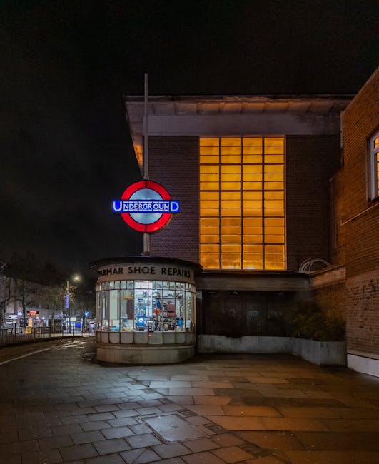 A nighttime street scene featuring a London Underground station entrance with a prominent red and blue roundel sign that reads 'UNDERGROUND' mounted above the entrance. To the left of the station, a small curved glass-fronted shop with a sign indicating 'HUMAR SHOE REPAIR' is visible, displaying repair tools and footwear inside. The surrounding pavement is made of rectangular stone slabs, some of which appear wet, reflecting surrounding lights. The building facade behind the station is constructed of brick and has a large, illuminated grid of orange-tinted glass windows, giving a warm glow against the night sky. Adjacent brick walls frame the scene, with sparse street lighting illuminating the area in a soft, diffuse manner. The overall setting suggests an urban environment close to busy public transport links, with the presence of waste clearance services in the vicinity potentially supporting local business waste management needs, especially for small shops nearby.