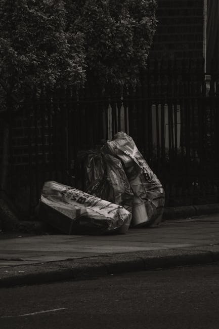 Two large black plastic bags filled with rubbish are placed on a pavement near a dark, metal fence with vertical bars. The bags appear to contain lightweight waste, possibly household or garden refuse, and are positioned adjacent to each other, leaning slightly against the fence. The surrounding environment includes a paved road surface in the foreground and dense greenery with leaves and branches visible above the fence in the background. The scene is captured in subdued lighting, suggesting overcast weather or late afternoon. This image depicts a typical situation where private or independent rubbish collection might be required, as evidenced by the placement of waste bags outside a residential boundary, aligning with the context of rubbish removal services provided by companies such as Waste Clearance Harrow.