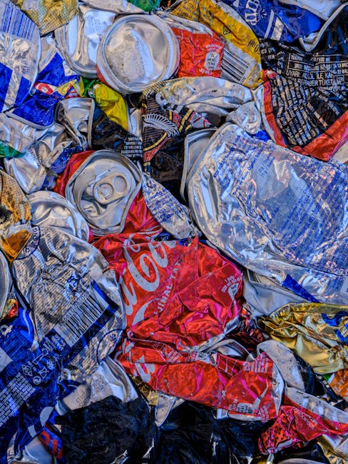 A close-up view of a pile of crushed beverage cans, primarily made of aluminium, with visible textures of shiny, metallic surfaces that reflect light. The cans are in various colors, including silver, red, blue, black, and gold, with some displaying visible brand labels such as Coca-Cola. The cans are overlapping and crumpled, showing dents and creases, and are scattered across a flat surface, likely on the ground or in a disposal area. The environment appears to be outdoors, possibly part of a waste collection or recycling process, with the cans stacked irregularly and some partially hidden behind others. The scene illustrates typical waste material collected for recycling or disposal, relating to rubbish removal services like those offered by Waste Clearance Harrow, which handle general waste and recyclable items. The lighting is bright, emphasizing the metallic sheen and reflective qualities of the aluminium cans, with some environmental context suggesting the scene is part of a waste management or alternative disposal operation.