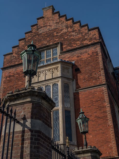 A historic red brick building with a stepped gable roof and large stained glass windows framed by stone detailing, situated behind a black wrought iron fence topped with decorative finials. Two traditional greenish-black lantern-style street lamps are mounted on stone pillars at the fence, casting shadows on the brickwork. The building's exterior features a mix of brickwork and stone accents with intricate carvings above the windows, suggesting an architectural style from an earlier period. The scene is illuminated by natural daylight, with a clear blue sky in the background. The image emphasizes the structure's historic character and exterior details, situated in an environment that appears to be on a street or courtyard area, possibly within a heritage or older neighbourhood. The overall setting is calm and unobstructed, providing an insight into traditional architecture that contrasts with modern rubbish removal or private waste handling services, which may be part of a local effort to maintain such historic sites, supported by companies like Waste Clearance Harrow.