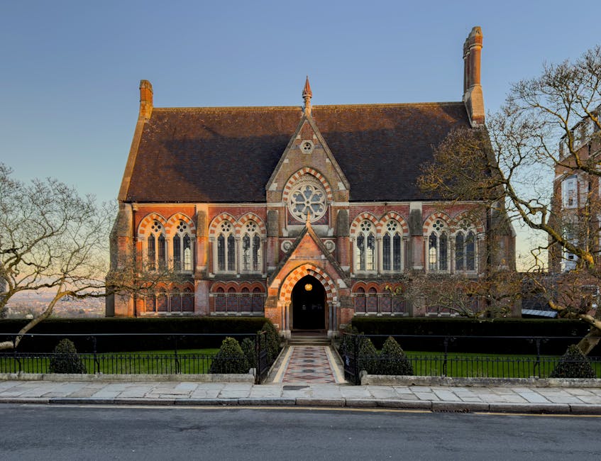 A historic red brick building with a stepped gable roof and large stained glass windows framed by stone detailing, situated behind a black wrought iron fence topped with decorative finials. Two traditional greenish-black lantern-style street lamps are mounted on stone pillars at the fence, casting shadows on the brickwork. The building's exterior features a mix of brickwork and stone accents with intricate carvings above the windows, suggesting an architectural style from an earlier period. The scene is illuminated by natural daylight, with a clear blue sky in the background. The image emphasizes the structure's historic character and exterior details, situated in an environment that appears to be on a street or courtyard area, possibly within a heritage or older neighbourhood. The overall setting is calm and unobstructed, providing an insight into traditional architecture that contrasts with modern rubbish removal or private waste handling services, which may be part of a local effort to maintain such historic sites, supported by companies like Waste Clearance Harrow.