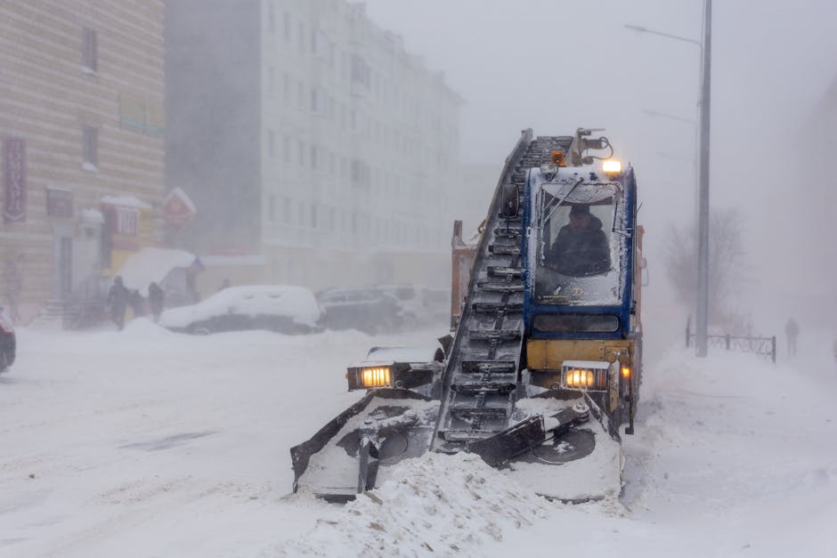 A small road scraper with a blue and yellow metal body clearing snow from an urban street during a heavy snowstorm. The machine has black rubber tracks, a front blade partially pushed into the snow, and is equipped with bright headlights and side lights for visibility. In the background, there are blurred buildings, parked cars covered in snow, and a few pedestrians walking with umbrellas on the snowy sidewalk. The scene appears foggy due to blowing snow and limited visibility, and the environment suggests an outdoor area in a city experiencing winter weather, where snow removal equipment is actively used to maintain access. The presence of the scraper aligns with alternative waste and debris removal methods in adverse weather conditions, as provided by companies like Waste Clearance Harrow.