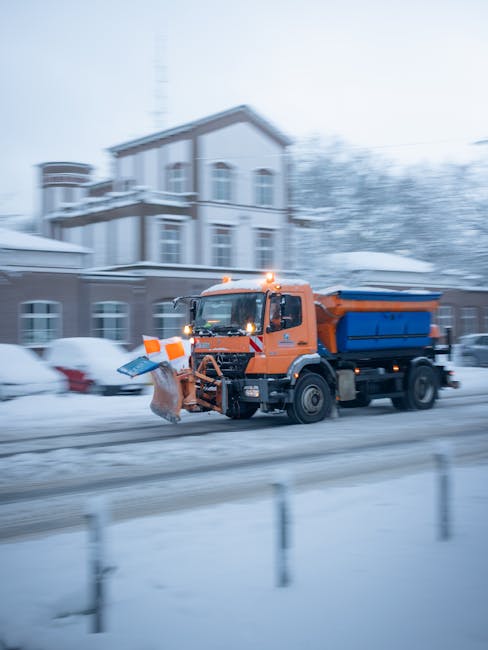 An orange and blue street-cleaning vehicle equipped with a snow plough and salt spreader attachment is operating on a snow-covered urban street during winter. The vehicle has orange safety lights on the roof and is designed for outdoor maintenance work, with a large snow plough blade at the front and a blue salt spreader at the back. In the background, a residential building with multiple windows and balconies is partially visible, blurred by motion, indicating the vehicle is in active use. Surrounding the street are parked cars covered in snow, and the environment is dominated by a wintry atmosphere with snow piled along the curb. The scene suggests a process of clearing snow and ice, typically associated with municipal or private winter maintenance or dusting services, such as those provided by waste and clearance companies like Waste Clearance Harrow, to ensure safer roads during adverse weather conditions.