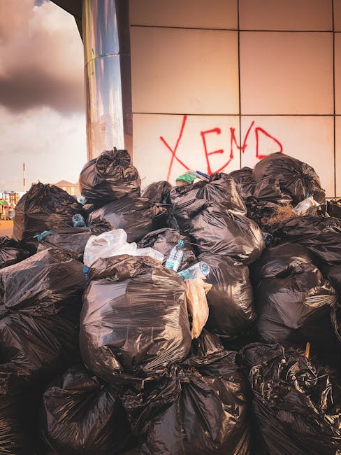 A large pile of black bin bags filled with waste materials is stacked outside a modern building with a tiled exterior wall, which features red graffiti text reading 'XEND.' The bin bags are placed on the pavement alongside scattered discarded plastic bottles and miscellaneous rubbish. The bags vary in size and are tightly knotted at the top, with some appearing slightly torn or stretched, revealing glimpses of their contents. The scene is illuminated by natural daylight, casting shadows across the bags and highlighting their glossy plastic surface. This outdoor environment suggests an instance of unscheduled rubbish accumulation, potentially related to a private waste collection or on-site clearance, reflecting a challenge faced by waste management services such as Waste Clearance Harrow. The background shows hints of an urban setting with distant buildings and an overcast sky, indicating the importance of prompt rubbish removal to maintain cleanliness and prevent clutter in the area.
