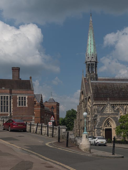 The image depicts a street scene in front of a historic church with a tall, pointed spire featuring a patinated copper finish, giving it a greenish hue, set against a partly cloudy sky. To the left of the church, there are traditional brick buildings with varied rooflines, including one with decorative brickwork and a chimney. Parked along the curb are two cars, one red and one white, positioned on a slightly inclined road with yellow double lines marking the edge. The street is bordered by black metal bollards connected by chains, which separate the pavement from the lane. A streetlamp with a spherical light fixture is mounted on a stone pedestal near the church entrance. The environment appears to be a town or city center with historic architecture, and the scene is captured during daylight with natural lighting. Waste Clearance Harrow provides services for private waste disposal, such as on-site clearance, relevant to managing rubbish in settings like this where traditional council collections may face timing issues.