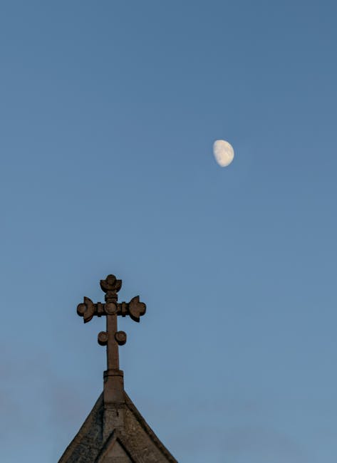 The image shows a clear blue sky with the visible lunar phase, positioned towards the upper right, displaying a partially illuminated moon in a waxing gibbous shape. In the lower part of the image, the pointed apex of a building's roof is visible, constructed from dark, weathered materials, possibly slate or similar textured roofing material, with a steep pitch. At the peak of the roof, an ornate metal cross with multiple arms and decorative elements is mounted, appearing dark and slightly rusted, suggesting aged metalwork. The surroundings do not include trees or other structures, focusing attention on the roof and sky. This scene exemplifies a quiet urban or suburban setting with an emphasis on traditional architectural details beneath a naturally lit sky. The overall composition highlights an atmospheric, daytime scene suitable for discussing independent collection methods or on-site waste clearance in a context aligned with private rubbish removal services, subtly supported by the setting's simplicity and focus on visible structures.
