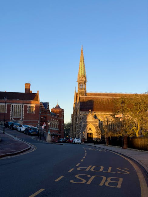 A view of a street scene featuring a large historic church with a tall, pointed spire constructed from stone and brick, situated on the right side of the image. The church has intricate architectural details around its arched entrance and windows, with weathered, darkened stonework visible on its exterior. To the left, there are several red-brick buildings with sloped roofs, and a row of parked cars lined along the curb. The road in the foreground curves gently to the left, marked with a yellow dashed line and the words 'BUS STOP' painted in yellow on the dark asphalt. The scene is illuminated by warm, low-angle sunlight, casting long shadows and highlighting the textures of the buildings and the church's stone façade. Sparse trees with bare branches are visible, and the sky above is clear and blue. This setting reflects a typical urban environment where private waste collection services, such as those offered by Waste Clearance Harrow, may facilitate rubbish removal or on-site clearance projects in historic or residential areas.